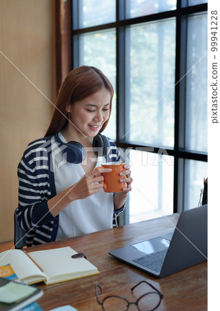 Portrait of a young, attractive Asian teenage girl using a computer and drinking coffee while studying online in the morning at the library. 99941228