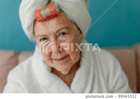 Happy senior woman with plastic curler sitting at sofa in bathrobes and looking at camera. 99943312