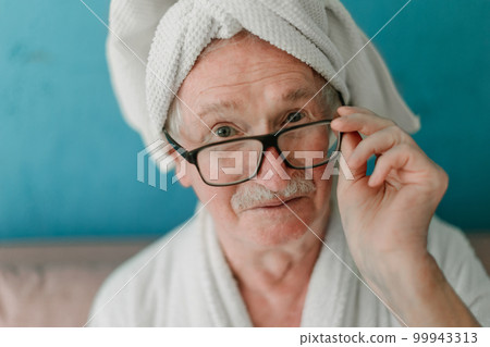 Happy senior man in eyeglasses sitting at sofa in bathrobes and looking at camera. 99943313