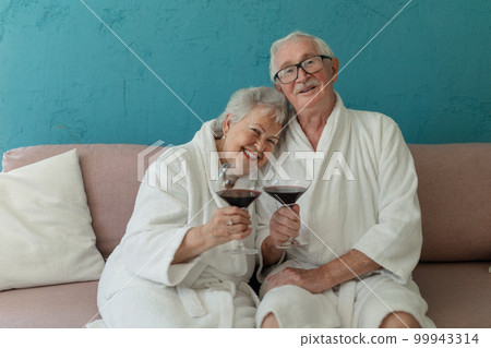 Happy senior couple sitting together in bathrobe on sofa with glass of wine, having nice time at home. 99943314