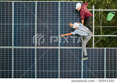 Aerial view of men technicians installing solar panels to high steel platform. Engineers wearing uniform and helmet. Concept of alternative energy. Aerial view of men technicians installing solar panels to high steel platform. Engineers wearing uniform and helmet. Concept of alternative energy. 99943388