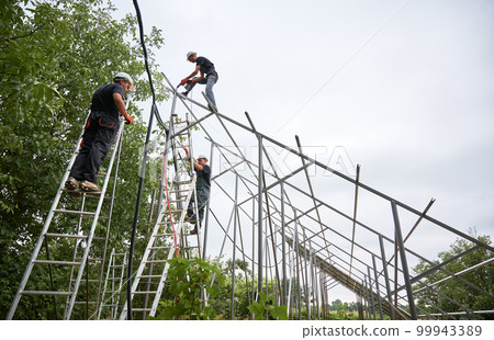 Three men in workwear installing metal poles and rails for photovoltaic solar panels. Male workers using ladders and drill while mounting support structures for solar panels. 99943389