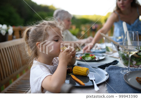 Family eating at barbecue party dinner on patio, little girl eating roasted corn and enjoying it. Family eating at barbecue party dinner on patio, little girl eating roasted corn and enjoying it. 99943390