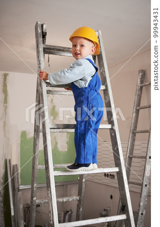 Child construction worker on the top of ladder while working on home renovation. Kid in safety helmet and work overalls walking up staircase in apartment under renovation. 99943431