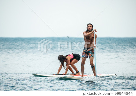 Sea woman and man on sup. Silhouette of happy young woman and man, surfing on SUP board, confident paddling through water surface. Idyllic sunset. Active lifestyle at sea or river. 99944844