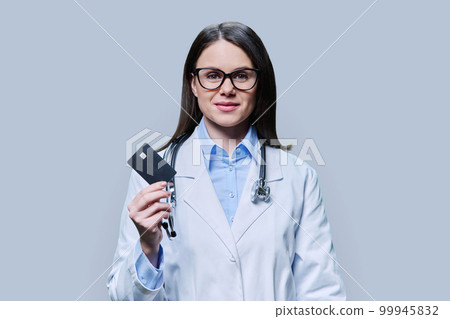 Young woman doctor holding credit card on light gray background Young woman doctor holding credit card on light gray background 99945832