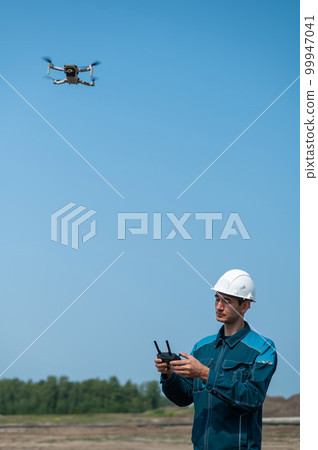 A man in a helmet and overalls controls a drone at a construction site. The builder carries out technical oversight. A man in a helmet and overalls controls a drone at a construction site. The builder carries out technical oversight. 99947041