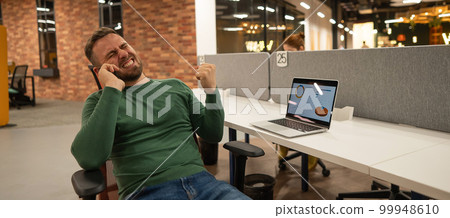 Caucasian bearded man talking on a smartphone while sitting at his desk in a modern coworking space. 99948610