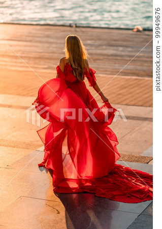 Sunrise red dress. A woman in a long red dress against the backdrop of sunrise, bright golden light of the sun's rays. The concept of femininity, harmony. 99949676