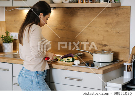Back view of female cutting different vegetables for preparing vegan dish at her kitchen 99951050