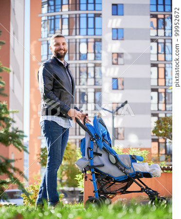 Loving father spending time with newborn kid outside. Side view of young bearded man in casual wear walking with baby stroller on street, with high-rise buildings on background. Concept of parenting. 99952672