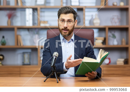 Senior mature pastor reading bible book online, man looking at camera sitting in home office and recording audio book online. Senior mature pastor reading bible book online, man looking at camera sitting in home office and recording audio book online. 99953068