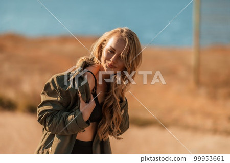 Portrait blonde sea cape. A calm young blonde in an unbuttoned khaki raincoat stands on the seashore, under the raincoat there is a black skirt and top 99953661