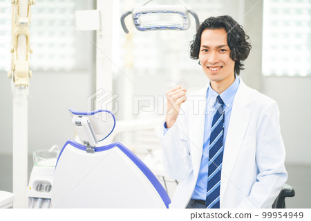 Young male dentist doing fist pose in front of dental treatment unit 99954949