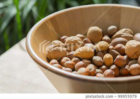Assorted various nuts in a big wooden bowl against a leafy green background Assorted various nuts in a big wooden bowl against a leafy green background 99955079