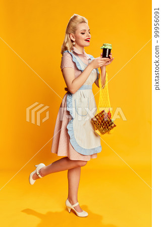 Food shopping. Portrait of beautiful young girl with stylish hairstyle posing against yellow studio background. Concept of beauty and pin-up style 99956091