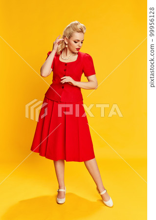 Femininity. Portrait of beautiful young blonde woman with stylish hairstyle in red suit posing against yellow studio background. Concept of beauty and pin-up style Femininity. Portrait of beautiful young blonde woman with stylish hairstyle in red suit posing against yellow studio background. Concept of beauty and pin-up style 99956128