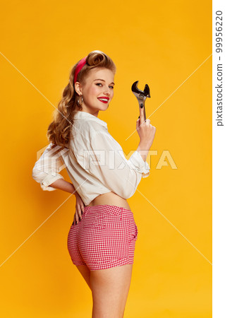 Portrait of beautiful young blonde girl in white shirt and pink shorts posing with tool, wrench against yellow studio background. Concept of beauty and pin-up style 99956220