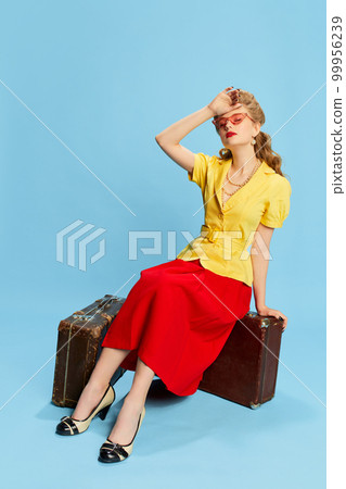 Feeling tired. Portrait of beautiful young girl in yellow blouse and red skirt sitting on vintage suitcases against blue studio background. Concept of beauty and pin-up style Feeling tired. Portrait of beautiful young girl in yellow blouse and red skirt sitting on vintage suitcases against blue studio background. Concept of beauty and pin-up style 99956239