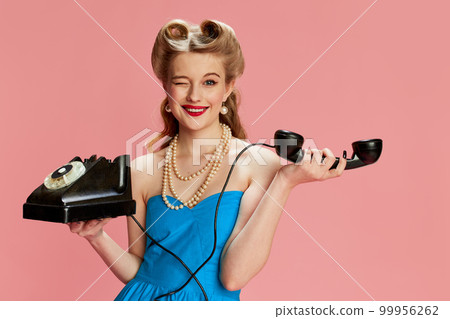 Portrait of beautiful young girl in blue dress and stylish hairstyle posing with vintage phone against pink studio background. Concept of beauty and pin-up style 99956262