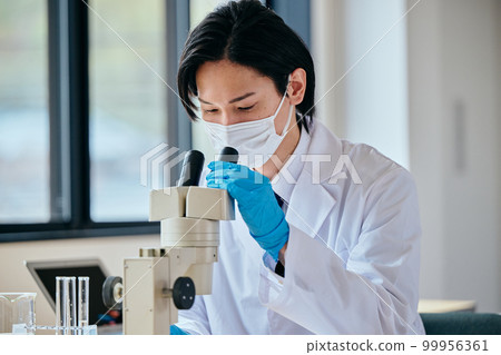 A young man in a white coat looking through the lens of a microscope and observing 99956361