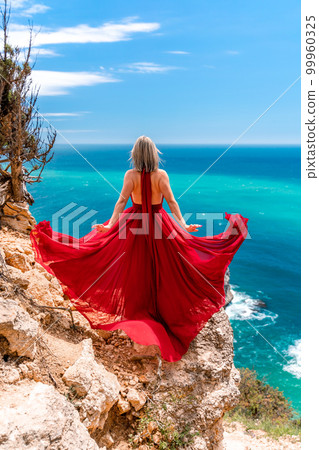 A girl with loose hair in a long red dress descends the stairs between the yellow rocks overlooking the sea. A rock can be seen in the sea. Sunny path on the sea from the rising sun A girl with loose hair in a long red dress descends the stairs between the yellow rocks overlooking the sea. A rock can be seen in the sea. Sunny path on the sea from the rising sun 99960325