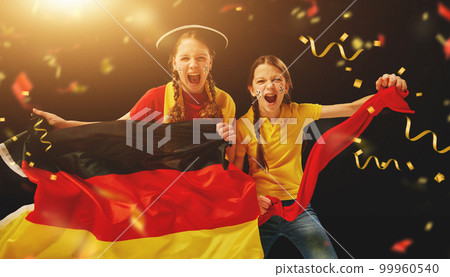 Young girls emotionally watching football match, holding flag and cheering team over dark background with confetti 99960540