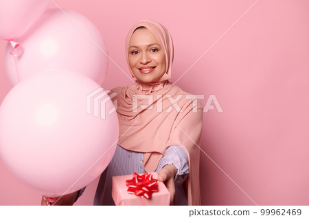 Fashion smiling Middle-Eastern Muslim woman holding pastel air balloons and a cute gift box, wearing hijab, looking at camera, isolated on pink color background. Arab female celebrating birthday party Fashion smiling Middle-Eastern Muslim woman holding pastel air balloons and a cute gift box, wearing hijab, looking at camera, isolated on pink color background. Arab female celebrating birthday party 99962469