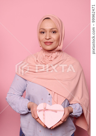 Attractive confident Middle-Eastern Muslim woman in pink hijab, showing at camera a heart shaped gift box on isolated background. Happy Mother's Day. Women's Day. Celebrating festive event, occasion Attractive confident Middle-Eastern Muslim woman in pink hijab, showing at camera a heart shaped gift box on isolated background. Happy Mother's Day. Women's Day. Celebrating festive event, occasion 99962471
