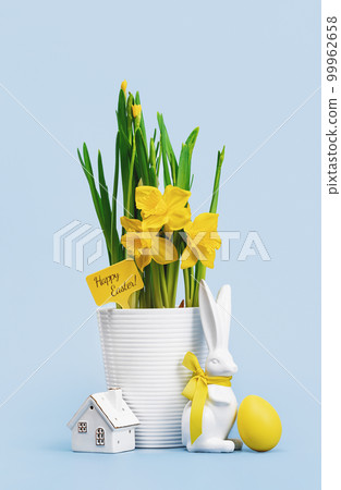 Yellow Easter egg and blooming daffodils in a white pot and Easter bunny with tiny house on blue background. tag with Happy Easter text. tall card 99962658