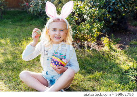 Portrait of happy little girl wearing bunny ears holding Easter egg after outdoor festive egg hunting in the garden. Kid having fun on Easter holiday. Children searching for eggs. Selective focus 99962796