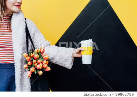 Cropped woman holding reusable coffee cup and fresh tulips in net bag on the geometry bright yellow and black wall background. Urban city street fashion. Spring mood. Coffee time. Selective focus. 99962799