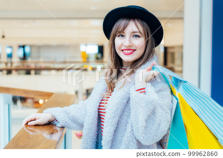 Smiling young fashion woman with net bag with fresh tulips flowers and paper shopping bags on mall background. Woman enjoying new spring season shopping. Consumerism, shopping, sale concept. Smiling young fashion woman with net bag with fresh tulips flowers and paper shopping bags on mall background. Woman enjoying new spring season shopping. Consumerism, shopping, sale concept. 99962860