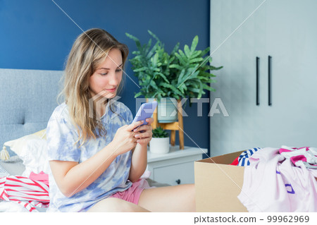 Smiling woman using smartphone to find places where to donate or sale clothes. Decluttering, Sorting clothes and Cleaning Up. Conscious consumer, sustainability. Reuse, second hand, Charity concept 99962969
