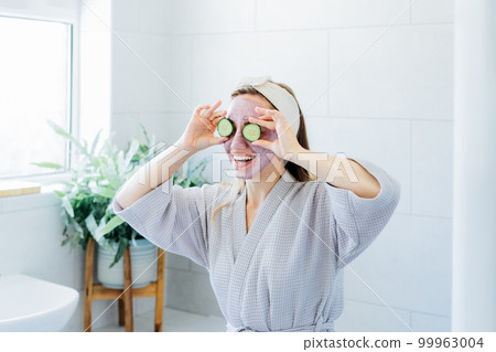 A young smiling woman with pink clay facial mask holds cucumber slices making a refreshing eye mask in bathroom. Natural cosmetic procedures for skin care at home. Beauty self-care. Selective focus A young smiling woman with pink clay facial mask holds cucumber slices making a refreshing eye mask in bathroom. Natural cosmetic procedures for skin care at home. Beauty self-care. Selective focus 99963004