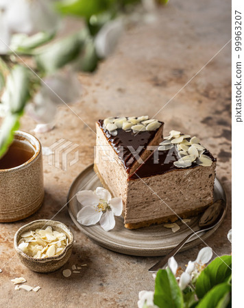 Two slices of chocolate cheesecake with almond petals, tea cup on brown textured background. Spring flowers in blur. Menu 99963207