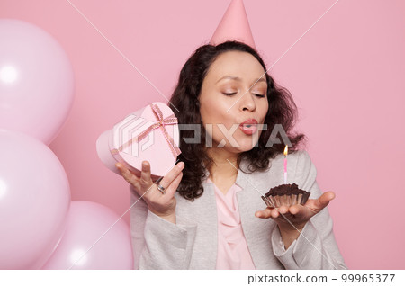 Sexy multi-ethnic middle-aged brunette woman, wearing a party hat, posing with pink pastel balloons, holding a gift box, blowing out a candle on birthday a cupcake, isolated on pink color background. 99965377