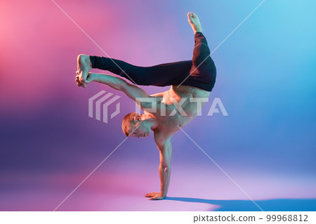 Young handsome acrobat or gymnast training to keep his body balance, standing on one hand while being barefoot and topless, touching his leg, showing his power. Young handsome acrobat or gymnast training to keep his body balance, standing on one hand while being barefoot and topless, touching his leg, showing his power. 99968812
