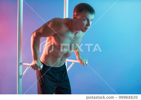 Young attractive man pulling up topless isolated over neon background, wearing black pants, looking straight ahead, exercising for his biceps, keeps fit. Young attractive man pulling up topless isolated over neon background, wearing black pants, looking straight ahead, exercising for his biceps, keeps fit. 99968820
