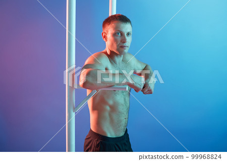 Handsome young man doing exercise on horizontal bar, guy posing topless, keeps fit, posing isolated over neon background, male looking aside with calm expression. Handsome young man doing exercise on horizontal bar, guy posing topless, keeps fit, posing isolated over neon background, male looking aside with calm expression. 99968824