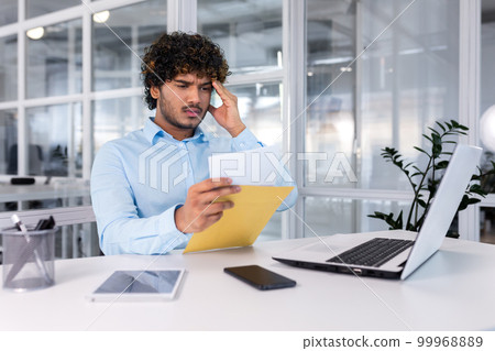 Young businessman at work inside office received mail envelope notification message with bad news, hispanic man sad reading letter document sitting at table with laptop. 99968889