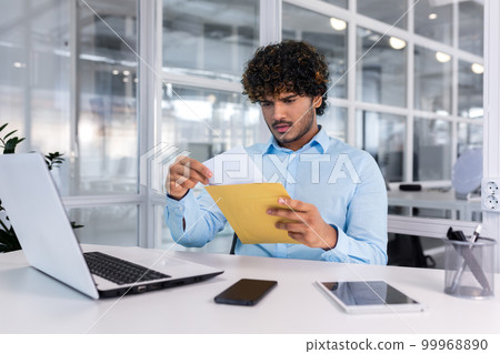 Young businessman at work inside office received mail envelope notification message with bad news, hispanic man sad reading letter document sitting at table with laptop. 99968890