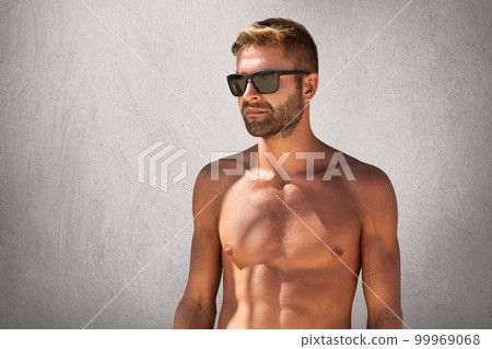 Waist up portrait of attractive macho man with bristle in trendy eyewear, standing naked against grey blank wall, looking aside with confident expression, demonstrating his suntanned, sport body Waist up portrait of attractive macho man with bristle in trendy eyewear, standing naked against grey blank wall, looking aside with confident expression, demonstrating his suntanned, sport body 99969068