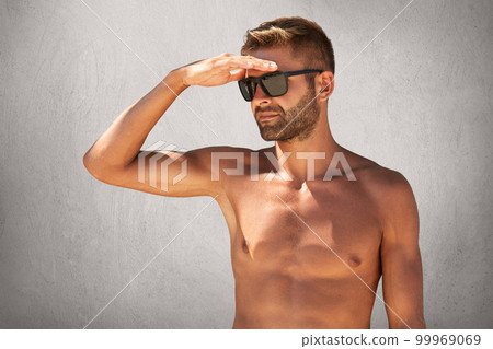 Portrait of handsome unshaven naked man in sunglasses, keeping his hand on forehead, looking into distance posing against grey concrete wall. Strong sportsman having summer vacations going to swim 99969069