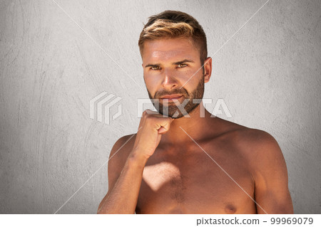 Serious man with bristle, dark attractive eyes, stylish hairdo standing topless while keeping his hand under chin isolated over grey background. Young Caucasian man with ideal musclular body 99969079