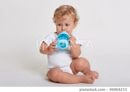 Baby drinking from baby cup while sitting on floor and looking away, wearing body suit, posing isolated over white background, male kid feels thirsty. Baby drinking from baby cup while sitting on floor and looking away, wearing body suit, posing isolated over white background, male kid feels thirsty. 99969233