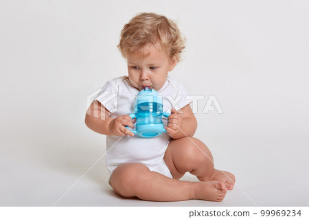 Little cute concentrated kid looking away while drinking water from baby cup, child with blonde curly hair sitting on floor, wearing body suit, posing isolated over white background. Little cute concentrated kid looking away while drinking water from baby cup, child with blonde curly hair sitting on floor, wearing body suit, posing isolated over white background. 99969234