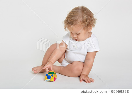 Tot with blond wavy hair playing with toy car while sitting barefoot on floor, child wearing white bodysuit looking at plastic toy, discovering new interesting thing. 99969258
