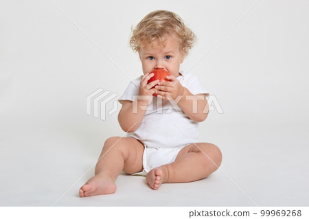 Beautiful blond curly haired little boy eats fresh red apple, healthy breakfast, starts day with healthy food, infant wearing body suit sitting barefoot on floor against white wall. Beautiful blond curly haired little boy eats fresh red apple, healthy breakfast, starts day with healthy food, infant wearing body suit sitting barefoot on floor against white wall. 99969268
