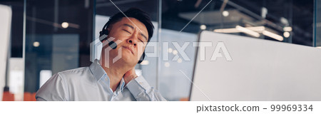 Tired asian businessman in headphones working on computer while sitting in office Tired asian businessman in headphones working on computer while sitting in office 99969334